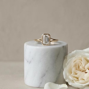 May include: A gold ring featuring an emerald-cut diamond. The diamond is set within a bezel. The ring is presented on a white marble cylinder, with a white rose in the background.