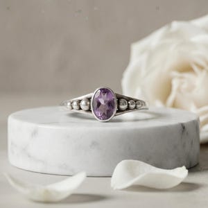 May include: A silver ring featuring a central oval-cut purple amethyst gemstone. The band is accented with three small silver spheres on each side of the stone, creating a symmetrical design. The metal has a polished finish with subtle dark oxidation.