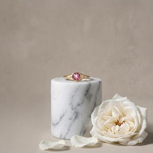 May include: A gold ring with a large, round, pink gemstone at the center, set on a white marble cylinder. The ring has intricate detailing and is displayed with a white rose and petals.