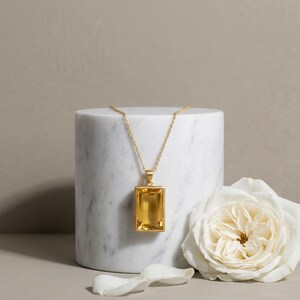 May include: A gold necklace featuring a rectangular, faceted yellow citrine gemstone pendant suspended from a delicate gold chain. The pendant has a polished gold frame that secures the transparent, honey-colored stone in a vertical orientation.