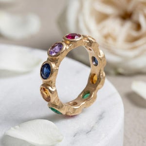 May include: A gold ring with a textured band, set with a sequence of colourful gemstones. The ring features oval-shaped gems in red, purple, blue, yellow, and green. The ring is displayed on a white marble surface, with a blurred floral background.