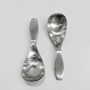 May include: Two polished stainless steel rice paddles with wide, rounded bowls and smooth, elongated handles. The metal surfaces feature a reflective, metallic finish with subtle contours, designed for serving cooked rice or other food items.