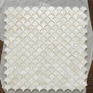 May include: A square sheet of iridescent, off-white mosaic tiles. The tiles are arranged in a fan or scallop pattern, creating a textured surface. The tiles are on a sheet of paper.