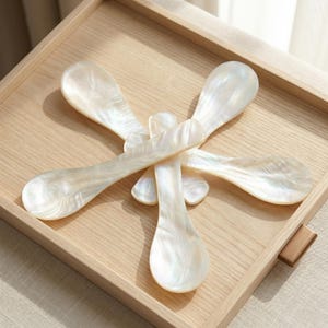 Puede incluir: Un juego de cinco cucharas nacaradas e iridiscentes dispuestas sobre una bandeja de madera clara. Las cucharas tienen un cuenco en forma de lágrima y un mango largo y delgado. La bandeja tiene un marco de madera marrón claro.