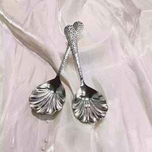Op de afbeelding: Twee zilverkleurige metalen lepels met kommen in de vorm van sint-jakobsschelpen. De handvatten hebben een getextureerde, gehamerde afwerking die uitloopt in een hartvormige top. Eén lepelkom bevat twee kleine, ronde, witte parelachtige accenten.