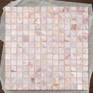 May include: A square mosaic tile sheet composed of numerous small, individual square tiles made of iridescent mother-of-pearl. The tiles feature a soft, light pink and white color palette with a natural, shimmering, and pearlescent finish.