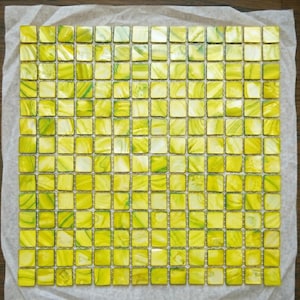 May include: A square sheet of small, square tiles in shades of yellow and green. The tiles have a glossy finish and are arranged in a grid pattern. The tiles are on a sheet of paper.