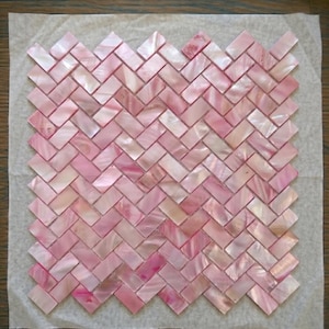 Op de afbeelding: Roze parelmoer mozaïektegels in een visgraatpatroon. De tegels variëren in roze tinten, wat een glinsterend effect creëert. De tegels liggen op een vel papier.
