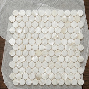 May include: A square sheet of white, round, mother-of-pearl mosaic tiles. The tiles are arranged in a grid pattern, with each tile slightly overlapping the next. The surface has a subtle, iridescent sheen, reflecting light. The tiles are on a sheet of paper.