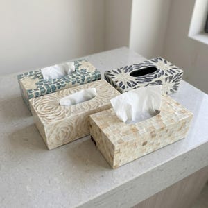 Puede incluir: Cuatro cajas rectangulares para pañuelos con distintos patrones decorativos. Una tiene un diseño de mosaico azul y blanco, otra presenta motivos de hojas gris oscuro, una muestra patrones florales blancos tallados y otra una cuadrícula crema.