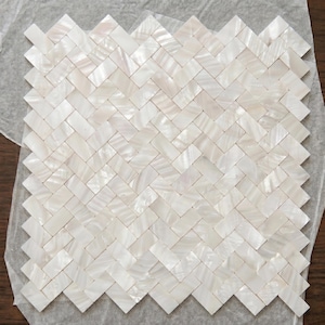 May include: A square mosaic tile sheet featuring small, rectangular, iridescent white tiles in a herringbone design. The tiles have a pearlescent sheen, reflecting light. The sheet is on a piece of paper.