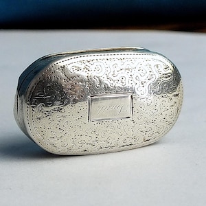 May include: A small, oval-shaped silver box with intricate engraved patterns and a rectangular cartouche. The box has a hinged lid and a decorative border. The word "Smith" is engraved on the cartouche.