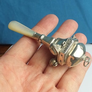 May include: A vintage silver baby rattle with a mother-of-pearl handle. The rattle features a decorative design with star and geometric accents. The overall length is approximately 10 cm.