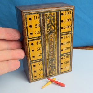 May include: A vintage wooden Bezique scoring box with a yellow and brown design. The box has numbered columns from 10 to 900, with the word "BEZIQUE" in the center. Two wooden pegs, one yellow and one red, are at the bottom.
