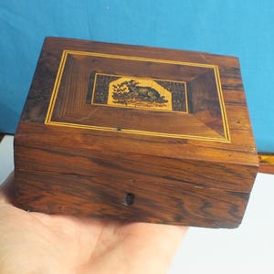Könnte beinhalten: Eine rechteckige Holzkiste mit dunkelbraunem Finish und eingelegten dekorativen Elementen. Die Kiste hat eine zentrale rechteckige Platte mit detailliertem Design, umrahmt von einer helleren Holzumrandung. Die Kiste wird in einer Hand gehalten.