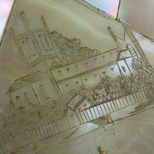 May include: A section of a mother-of-pearl inlay with an etched landscape scene. The scene depicts a building with multiple chimneys, trees, and a fence. The iridescent surface reflects light, creating a shimmering effect.