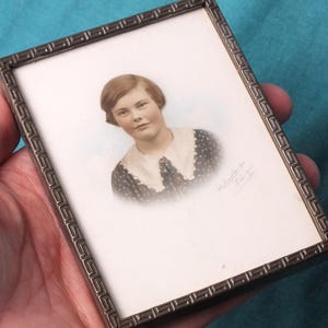 May include: A vintage portrait in a rectangular metal frame. The portrait features a woman with short brown hair, wearing a polka-dot dress with a white collar. The frame has a decorative border. The image is held in a hand.