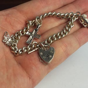 Può includere: Bracciale con ciondoli in argento con farfalla, croce, lucchetto a forma di cuore e una piccola auto. Il bracciale ha un design a catena spessa ed è realizzato in metallo color argento lucido. I ciondoli sono dettagliati.