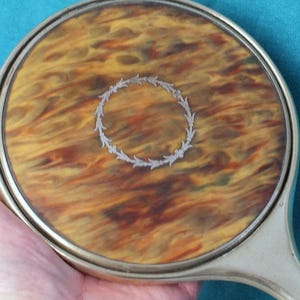 May include: A vintage hand mirror featuring a round face with a swirling pattern in brown, yellow, and orange hues. The mirror is framed in silver-coloured metal, with a decorative wreath in the centre. The handle is also silver-coloured.