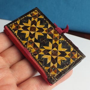 May include: A small rectangular box with a hinged lid featuring an intricate geometric mosaic pattern in shades of yellow, brown, and black. The sides of the box are covered in a vibrant red fabric, and a small red fabric tab is attached to the lid.