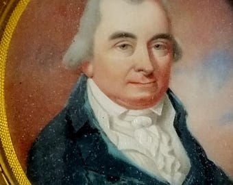 May include: Oval portrait miniature of a man with powdered white hair, wearing a dark blue coat and a white ruffled shirt. The portrait is set within a gold-colored frame, with a reddish-brown background.
