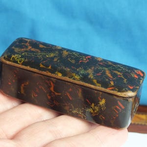 May include: A rectangular, antique box with a marbled pattern in shades of black, brown, red, and yellow. The box is held in a hand, showcasing its compact size and intricate design. The box has a gold-colored trim.