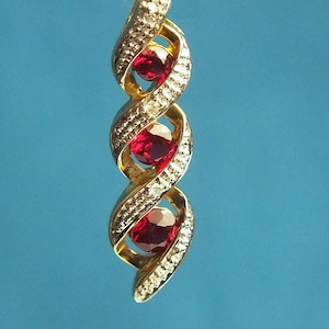 Pode incluir: Um pingente de ouro com um design de espiral vertical cravejado com três pedras preciosas redondas de cor vermelho profundo. A estrutura de metal em espiral é acentuada com pequenas pedras claras em pavé ao longo das bordas da peça.