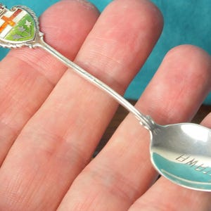 Op de afbeelding: Een zilveren souvenirlepel met een gedetailleerd emaille wapen met een kroon, kruis en schild met groene, oranje en witte accenten. De lepelbak is gegraveerd met het woord "OTTAWA".