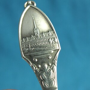 May include: A silver souvenir spoon with a detailed engraved image of a building with a tall spire. The spoon's handle features an ornate floral design. The spoon is made of a shiny, reflective metal.