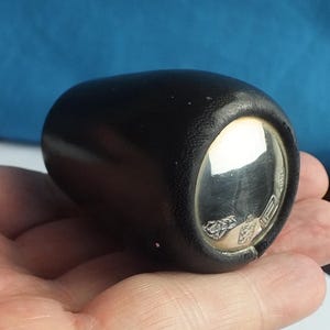 Puede incluir: Objeto cilíndrico negro con base plateada y reflectante. El objeto se sostiene en una mano, con la base plateada reflejando la luz. Parece ser un artículo decorativo o un pequeño recipiente.