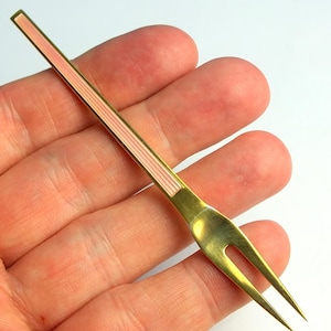 Puede incluir: Un pequeño tenedor de cóctel de metal dorado con un mango delgado y una incrustación vertical de esmalte rosa pálido. El tenedor tiene dos dientes afilados y un acabado pulido, mostrando un diseño sencillo con un tallo estrecho.