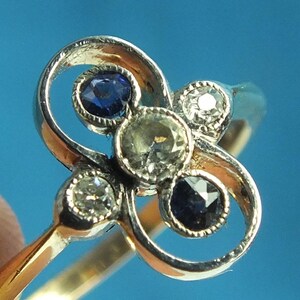 May include: A gold ring with a silver-colored floral design. The ring features five gemstones: three clear diamonds and two dark blue sapphires. The ring is set against a blue background.