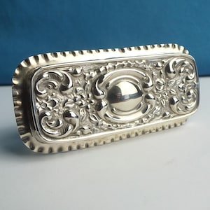 Puede incluir: Una caja rectangular plateada con detalles ornamentales de flores y volutas. La caja tiene un borde festoneado y un diseño ovalado central en relieve. La superficie es muy reflectante, lo que sugiere un acabado metálico pulido. El diseño es clásico y elegante.