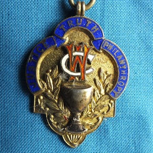 May include: Vintage gold-toned pendant with blue enamel text reading "TRUTH, JUSTICE, PHILANTHROPY." The pendant features a central emblem with the letters "WC" and a chalice. Laurel leaves and a small ring for hanging are also visible.