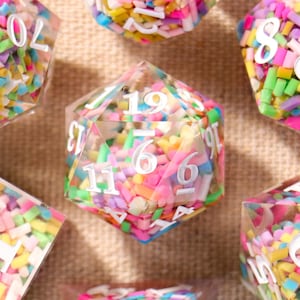 May include: A set of seven polyhedral dice. The clear resin dice are filled with colourful sprinkles and white numbers. The dice are in various geometric shapes, including a d4, d6, d8, d10, d12, and d20, ideal for role-playing games.