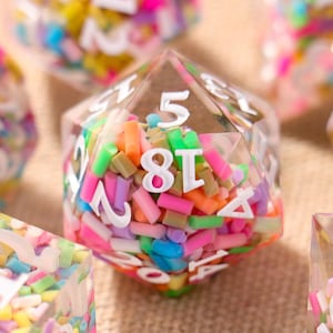 May include: Close-up of several polyhedral dice. The dice are clear with colourful sprinkles inside and white numbers. The dice are in various shapes, including a 20-sided die. The dice are on a neutral-coloured surface.
