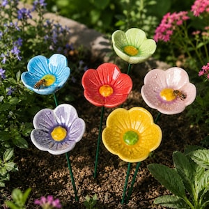 Könnte beinhalten: Eine Sammlung farbenfroher Gartenstecker in Form von Blumen aus Keramik. Jede Blume hat eine andere Farbe, darunter Blau, Rot, Gelb, Lila, Rosa und Grün, mit einem gelben oder grünen Zentrum. Einige haben eine Biene.