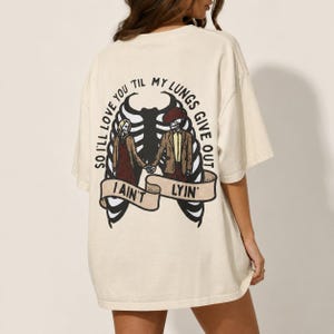 May include: An off-white short-sleeve t-shirt featuring a graphic on the back of two skeletons holding hands inside a ribcage. The text reads, "SO I'LL LOVE YOU 'TIL MY LUNGS GIVE OUT I AIN'T LYIN'" on a banner below the skeletons.