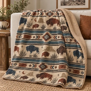 May include: A plush throw blanket featuring a cream-colored base with a repeating pattern of brown and blue bison, desert mesas, crescent moons, and stars, interspersed with geometric bands and a textured tan sherpa fleece lining on the reverse side.