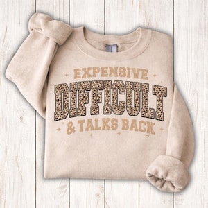 May include: A beige sweatshirt with the words "EXPENSIVE DIFFICULT & TALKS BACK" printed on the front. The word "DIFFICULT" is in a leopard print pattern. The sweatshirt has long sleeves and a crew neck.
