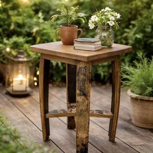 May include: A small, square wooden side table with a rustic barrel-stave design. The table holds a potted plant, a small stack of books, and a glass vase with white flowers. The table is on a wooden deck.