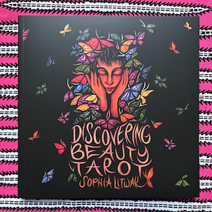 Peut inclure: Un jeu de cartes de tarot noir intitulé "Discovering Beauty Tarot" par Sophia Lütwak. La couverture présente une femme avec des papillons dans ses cheveux et autour d'elle, avec le titre en script orange et rouge.