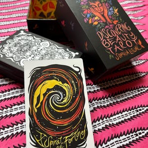 May include: A tarot card with a spiral design and the text "A Spiral of Fortune" is displayed in front of a black box set. The box set is labeled "Discovering Beauty Tarot" with an illustration of a woman's face surrounded by butterflies.