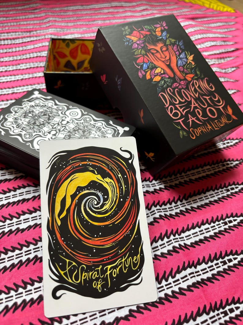 May include: A tarot card with a spiral design and the text "A Spiral of Fortune" is displayed in front of a black box set. The box set is labeled "Discovering Beauty Tarot" with an illustration of a woman's face surrounded by butterflies.