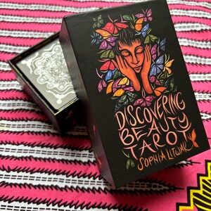 May include: Black tarot card box with the words "Discovering Beauty Tarot" and the author's name. The box features a colorful illustration of a woman's face surrounded by butterflies. The box is open, revealing the tarot cards inside.