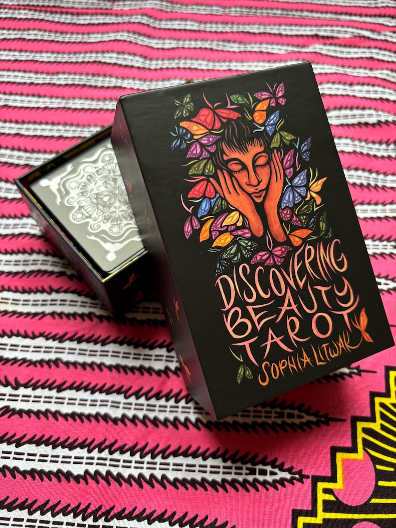May include: Black tarot card box with the words "Discovering Beauty Tarot" and the author's name. The box features a colorful illustration of a woman's face surrounded by butterflies. The box is open, revealing the tarot cards inside.
