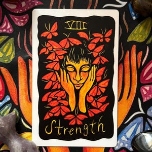 May include: A tarot card with a black background featuring a woman's face surrounded by orange leaves. The word "Strength" is written in yellow script below the image. The card is surrounded by colorful crystals and a patterned fabric.