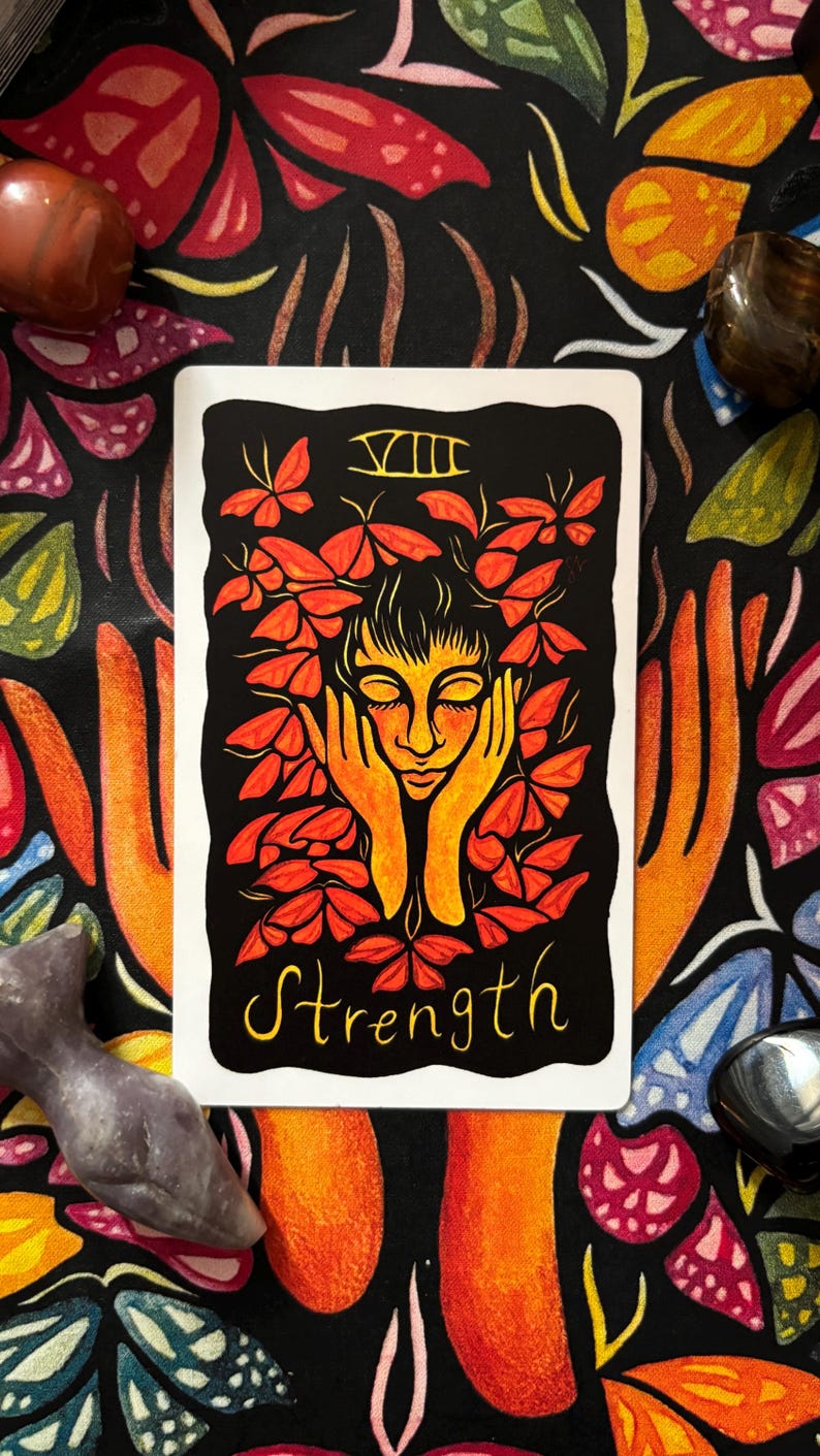 May include: A tarot card with a black background featuring a woman's face surrounded by orange leaves. The word "Strength" is written in yellow script below the image. The card is surrounded by colorful crystals and a patterned fabric.
