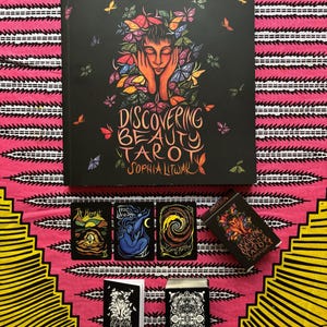 Peut inclure: Un jeu de tarot noir intitulé "Discovering Beauty Tarot" avec une illustration d'une personne avec des papillons. L'ensemble comprend un grand livre, trois cartes de tarot et deux petites boîtes, sur un fond rose et jaune.