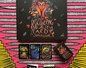 May include: A black tarot deck set with the title "Discovering Beauty Tarot" and artwork of a person with butterflies. The set includes a large book, three tarot cards, and two smaller boxes, all on a pink and yellow patterned background.